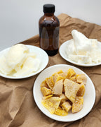 White dishes containing coconut oil, shea butter, and local raw yellow beeswax, alongside a bottle of fragrance oil. The makings for lotion bars.